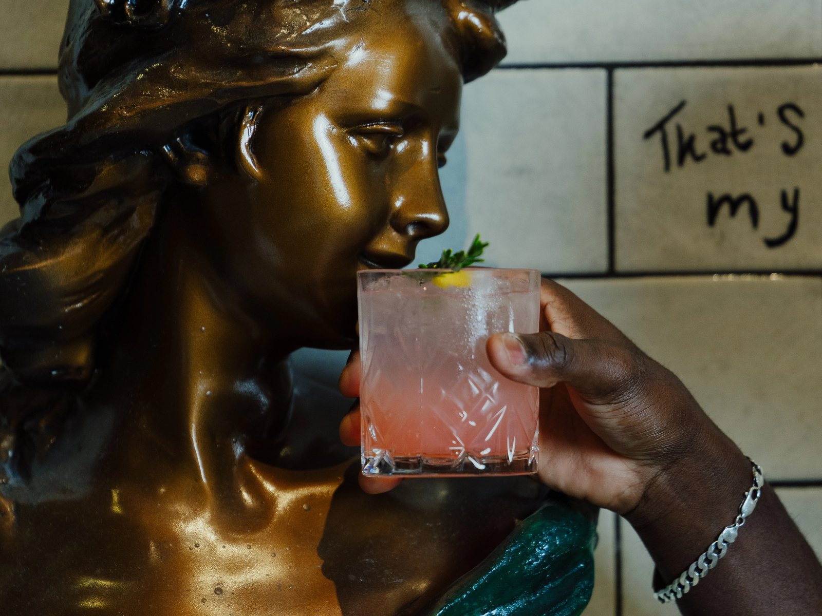 Statue de bronze du Conteur devant un mur de carrelage blanc, un cocktail à la main